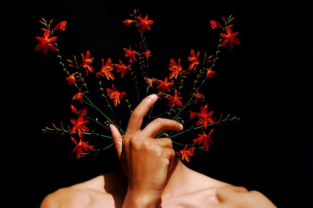 Main devant le visage d’un corps nu d’où émergent des fleurs rouges sur fond noir, symbolisant l’expression intérieure et la fusion entre l’humain et la nature.